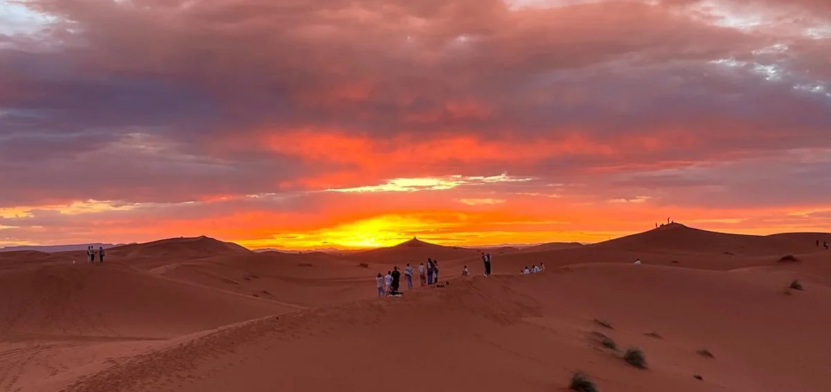 Sunset over the Erg Chebbi dunes in Merzouga with travelers walking on the Sahara Desert sand.